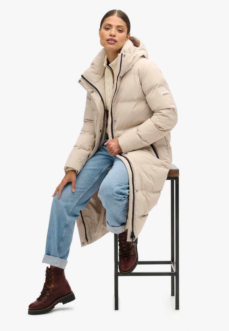 Beżowa kurtka puchowa z kapturem, z przednim zamkiem błyskawicznym i czarnymi akcentami, noszona na kremowym swetrze dzianinowym i jasnoniebieskich dżinsach, zestawiona z brązowymi botkami.