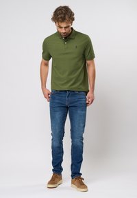T-shirt polo vert à manches courtes et col, assorti à un jean slim bleu et des baskets beiges, se tenant devant un fond neutre.