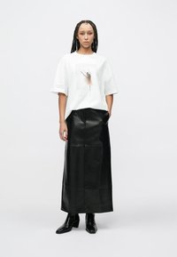 T-shirt branca oversized com uma estampa abstrata centrada, combinada com uma saia longa preta de couro e botas pretas com salto. Texturas lisas em todo o conjunto.