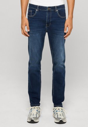 Homme portant un jean slim bleu foncé et des chaussures de sport blanches à lacets blancs, debout devant un fond uni.