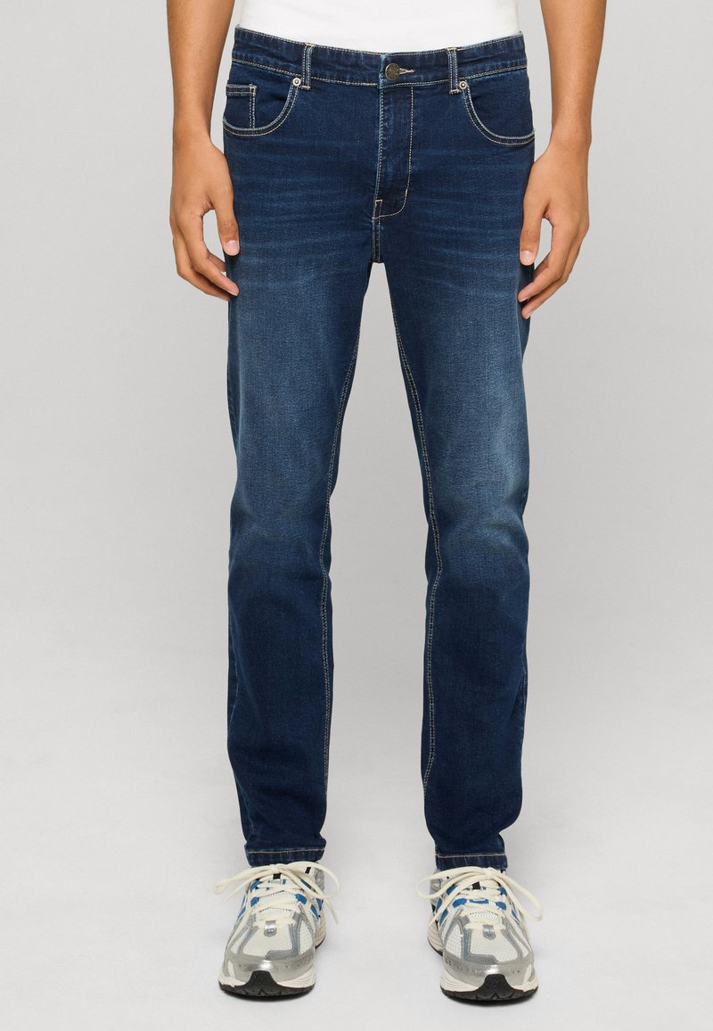 Homme portant un jean slim bleu foncé et des chaussures de sport blanches à lacets blancs, debout devant un fond uni.