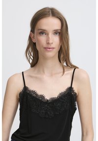 Junge Frau mit hellbraunem Haar, die ein schwarzes Top mit Spitze trägt, vor einem schlichten weißen Hintergrund.