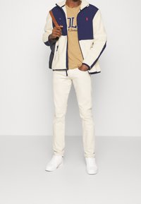 Bunda s fleecovou podšívkou v námořnické modři a krémové barvě, se zipem a kapsami. Pod ní béžové tričko, v kombinaci s bílými kalhotami a teniskami. Hnědá kabelka přes rameno.