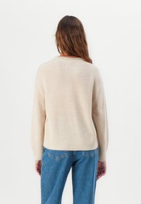 Pull en maille beige avec un col rond et une texture côtelée, doté de manches tombantes et d'une coupe décontractée, associé à un jean bleu.
