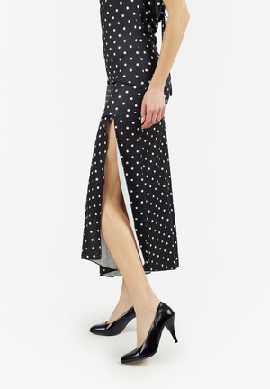 Donna che indossa una gonna nera a pois con uno spacco laterale alto e scarpe nere con tacco alto, in piedi su uno sfondo bianco.