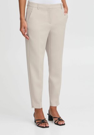 Nainen pukeutuneena beigenvärisiin mittatilaushousuihin, jotka ovat nilkkoihin ulottuvat ja joissa on sivutaskut, sekä mustat korolliset sandaalit, seisomassa yksiväristä taustaa vasten.