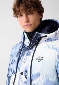 Veste rembourrée blanche et bleu clair avec un motif montagne, col haut, capuche à cordon de serrage et logo "Guess" brodé sur la poitrine.