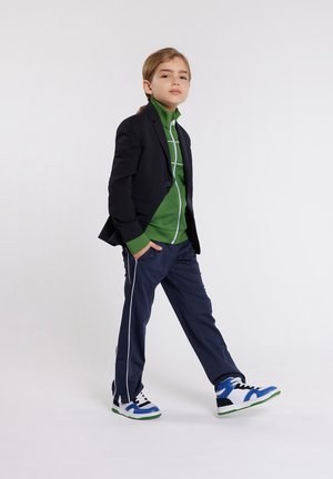 Blazer nero sopra un cappuccio verde con zip, abbinato a pantaloni sportivi blu navy con strisce bianche e sneakers blu con dettagli verdi.