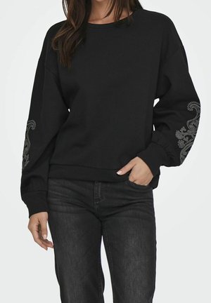 Femme portant un sweat-shirt noir à manches longues avec des motifs floraux gris sur les manches et un jean noir, une main dans la poche, sur fond blanc.