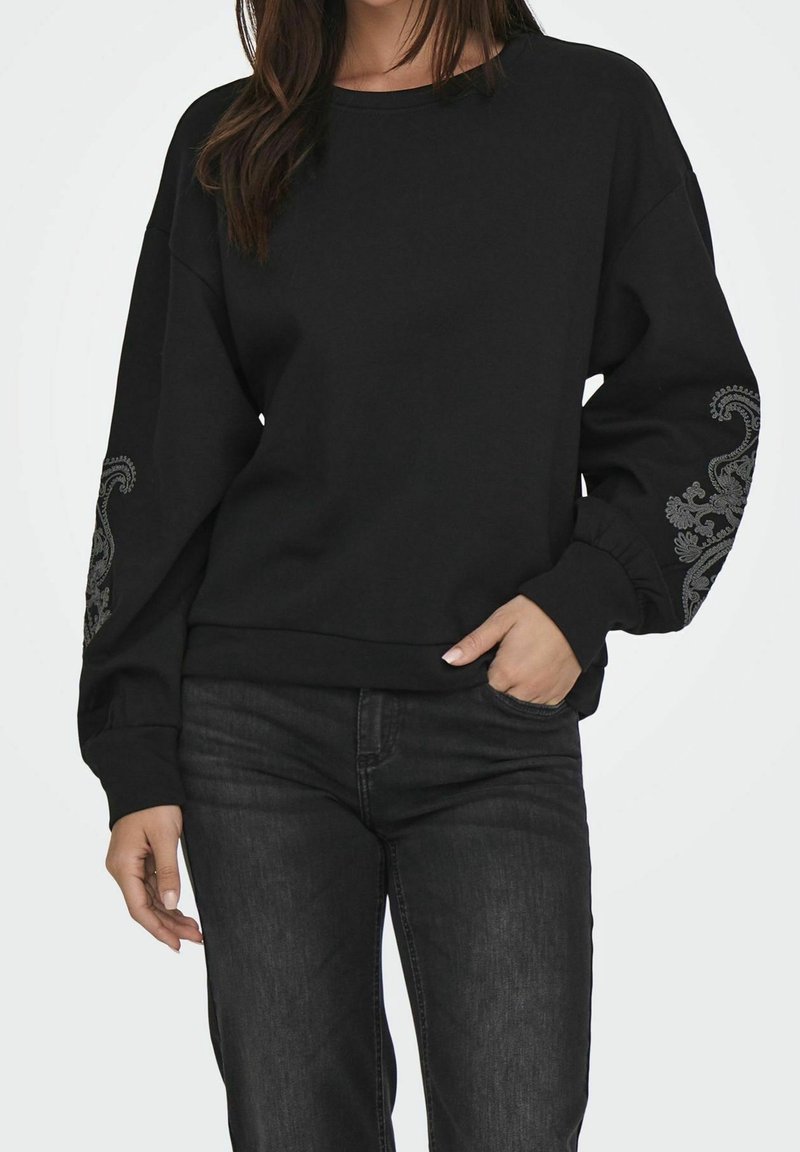 Femme portant un sweat-shirt noir à manches longues avec des motifs floraux gris sur les manches et un jean noir, une main dans la poche, sur fond blanc.