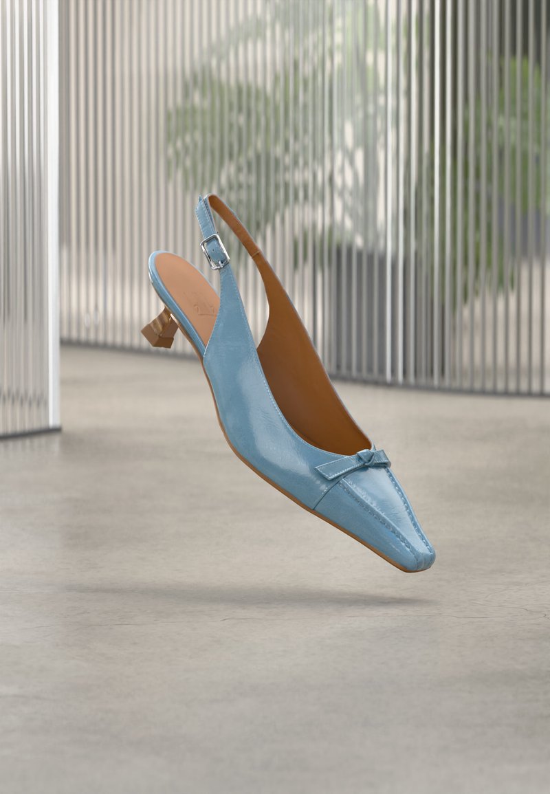Chaussure slingback en cuir verni bleu clair avec petit talon carré et nœud, flottant au-dessus d'un sol poli avec du verre côtelé et des plantes en arrière-plan.