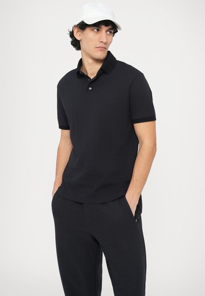 Fekete galléros póló texturált anyagból, gallérral, rövid ujjakkal és háromgombos gombolattal, hozzáillő fekete nadrággal párosítva.