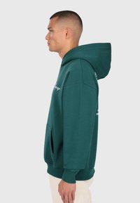 Mann in grüner Kapuzenjacke mit Fronttasche und cremefarbenen Hosen, im Profil vor einem einfarbigen hellen Hintergrund stehend.