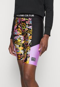 Barevné potisknuté cyklistické šortky mají květinový vzor na černém pozadí s růžovým panelem. Elastický pas zobrazuje logo "VERSACE JEANS COUTURE".