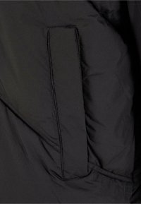 Schwarze Daunenjacke aus Stoff mit einer Seitentasche, die über gestickte Nähte und eine glatte, matte Textur verfügt.