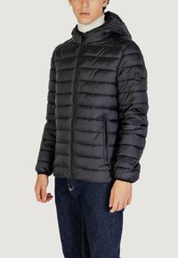 Chaqueta acolchada negra con capucha, que presenta un diseño de acolchado horizontal, bolsillos con cremallera y puños elásticos. Acompañada de pantalones de mezclilla oscuros.