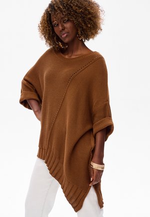 Vrouw met krullend haar, grote hoepeloorbellen, een losbruine gebreide poncho, witte broek en gouden armbanden om haar pols.