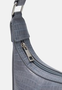 Torebka z szarej syntetycznej skóry o strukturze krokodylej, z detalem w postaci srebrnego suwaka i szerokim mocowaniem pasków, prezentująca zakrzywiony kształt.