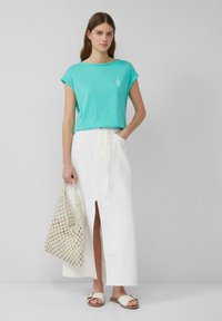 Turkusowa bawełniana koszulka z podwiniętymi rękawami, zestawiona z białą lnianą spódnicą z bocznym rozcięciem. Uzupełniona plecioną torbą.
