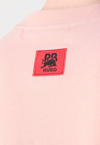 T-shirt en coton rose avec une étiquette rectangulaire rouge portant un logo noir et un design de taureau, située sur le haut du dos.
