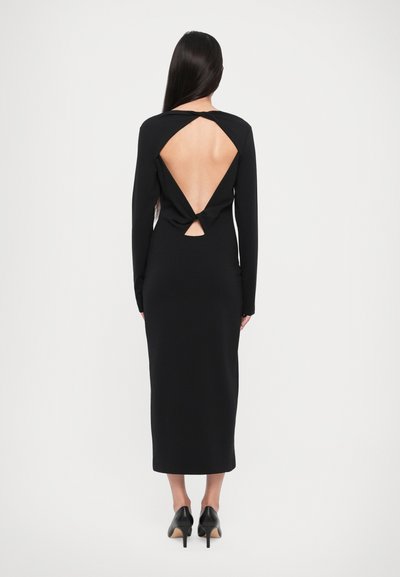 Black long-sleeve dress featuring an open back with knot detail, fitted silhouette, and mid-calf length, paired with black high-heeled shoes.