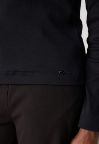 Marineblauwe top met lange mouwen, geribbelde structuur, ronde halslijn en een klein metalen accent met logo bij de zoom, gecombineerd met donkere broek.