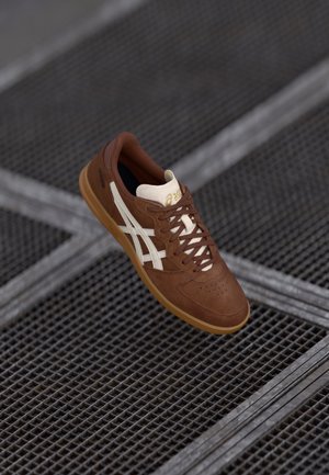 Brown and white Asics sneaker with laces, placed on a metal grid floor with intersecting lines in a diagonal pattern.