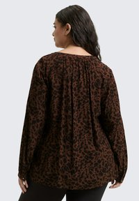 Woman with long dark hair wears a loose brown blouse with black leopard print, shown from the back against a plain light background.