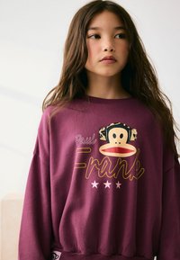 Hanorac purpuriu dintr-un material moale, decorat cu un grafic al unei fețe de maimuță cu urechi din imprimeu leopard și text galben. Include tiv elastic.