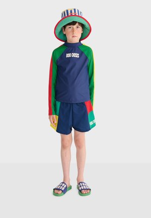 Dječak odjeven u šarenu dugomisnu majicu za kupanje, odgovarajuće kratke hlače s natpisom "BOBO CHOSES", prugasti šešir s obodom i plavo-zelene sandale na remen.