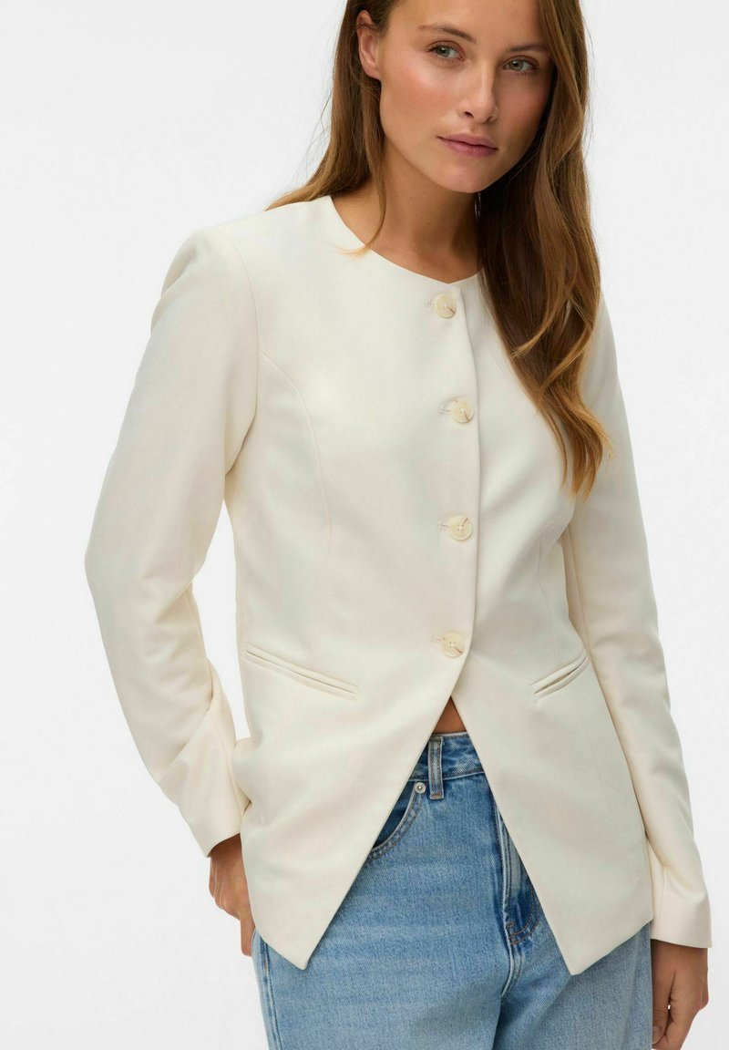 Femme portant un blazer crème ajusté à boutons avec une fente à l'avant et un jean bleu, debout les mains partiellement dans les poches.