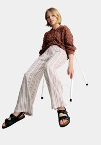 Pull en maille texturée marron, pantalon large rayé en crème et rose pâle, et sandales noires avec détails de boucle. Modèle assis sur une chaise.