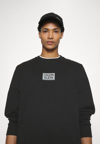 Felpa nera con logo "CALVIN KLEIN" grigio, scollo rotondo e polsini a coste. Cappellino nero con un accento frontale e visiera strutturata.