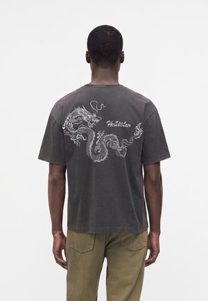 Uomo che indossa una t-shirt nera scolorita con una grafica dettagliata di un drago bianco e testo "Hollister" sulla schiena, abbinata a pantaloni verde oliva.
