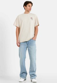 Camiseta de manga corta beige con un gráfico en la parte delantera, combinada con jeans rectos de color azul claro. Calzado casual en blanco completa el look.