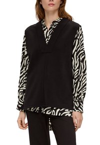 Top senza maniche nero con scollatura a V indossato sopra una camicia a maniche lunghe bianca e nera con stampa zebra. L'outfit è completato da pantaloni neri.