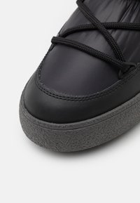 Schwarze Sneaker mit einem glänzenden Lederoberteil und einer strukturierten grauen Gummisohle. Merkmale sind schwarze Schnürdetails und ein abgerundetes Design der Zehenpartie.