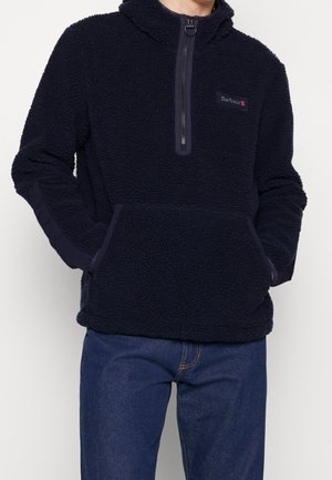 Mężczyzna ubrany w czarny polar z przednim zamkiem błyskawicznym i kieszenią typu kangur, w połączeniu z niebieskimi dżinsami, ręce w kieszeniach.