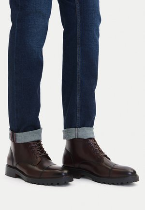 Brązowe skórzane botki z zaokrąglonym noskiem, wiązane na sznurowadła, z teksturowaną gumową podeszwą, zestawione z ciemnymi dżinsami, podwiniętymi na mankietach.