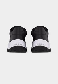 Scarpe da ginnastica chunky in bianco e nero con suole spesse, viste da dietro, con il marchio "GUESS" sulle suole intermedie bianche.