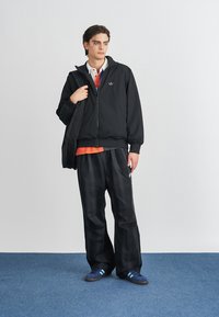 Crna jakna na rajsferšlus s kapuljačom, narančasta majica, karirani crni hlače i plave tene sa prugama. Model drži crnu torbu. Jednostavna pozadina.