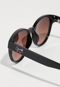 Occhiali da sole oversized neri con lenti sfumate marroni. Montatura robusta e design elegante con il logo "Guess" rosa sulla asta.
