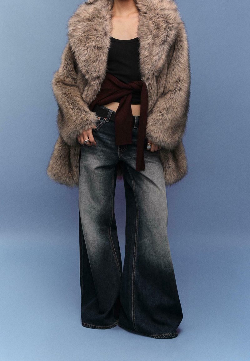 Personne portant un manteau en fourrure surdimensionné, un crop top noir, un pull noué autour de la taille et un jean large délavé sur fond bleu.