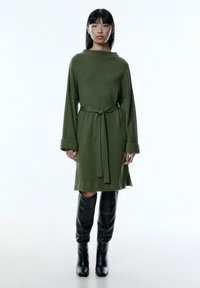 Olivengrünes, geripptes Strickkleid mit weitem Ausschnitt, tailliertem Bund und langen Ärmeln; kombiniert mit knielangen schwarzen Lederstiefeln.