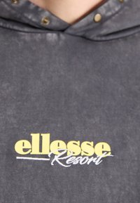 Sudadera gris con capucha ajustable con cordón, botones a presión y un logo estampado de "ellesse Resort" en amarillo y blanco. Textura suave.