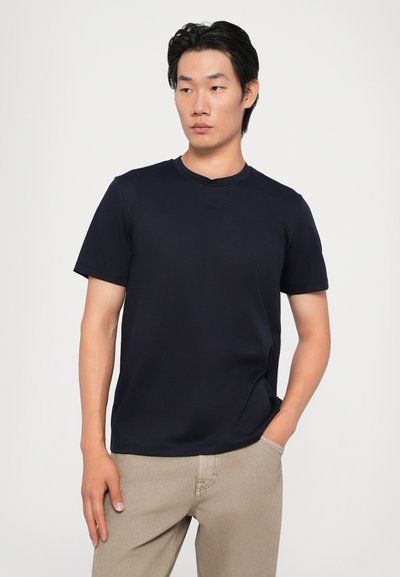 T-shirt bleu marine à manches courtes avec un col rond, en tissu lisse. Présente une coupe ajustée et un design minimaliste, porté avec un pantalon beige.