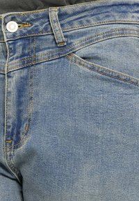 Κοντινή λήψη μπλε τζιν παντελονιών που δείχνει την εμπρόσθια τσέπη, τις θηλιές ζώνης, το μεταλλικό κουμπί και τις πορτοκαλί ραφές.