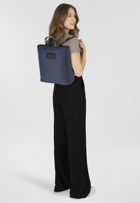 Mochila de nylon azul marino con asas superiores, forma cuadrada y un emblema en la parte frontal. Textura suave y diseño minimalista, llevada de manera casual.