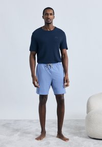 Camisa azul-marinho de manga curta com tecido texturizado, combinada com calções de banho azul claro com cordão ajustável. Postura descalço.