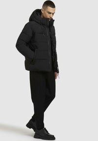 Schwarze Steppjacke mit Kapuze, ausgestattet mit einem gesteppten Design, einem Reißverschluss vorne und Seitentaschen. Getragen über schwarzen Hosen und Schuhen.